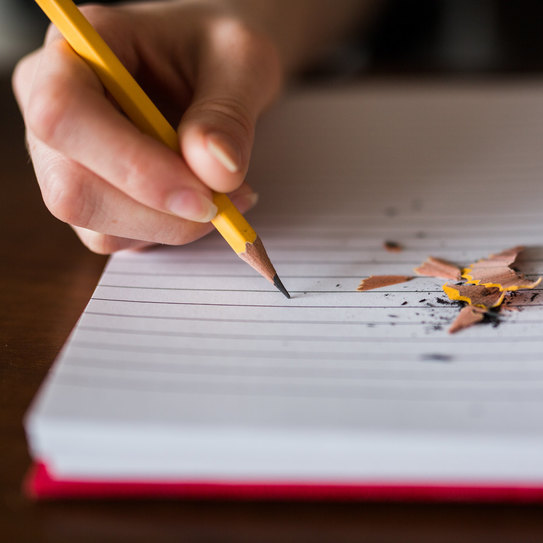 Das Foto ist die Nahaufnahme einer Hand, die mit Bleistift etwas in ein Notizbuch schreibt.