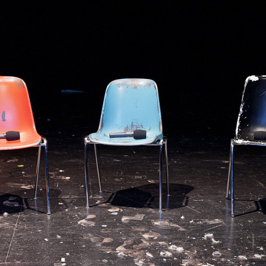 Das Foto zeigt drei Stühle auf einer Theaterbühne. Auf jedem Stuhl liegt ein Mikrophon.